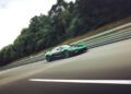 A green sports car drives at high speed on a highway, with trees and a metal barrier in the background.