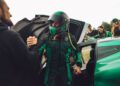 A person in a racing suit and helmet exits a green Rimac Nevera, the fastest EV that breaks records, shaking hands with another as a group of people look on.