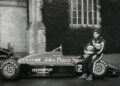 A race car driver in a racing suit sits on the front wheel of a John Player Special Formula 1 car, as Lotus prepares to honor F1 legends Senna and Clark at Goodwood, parked before a building with large windows and ivy-covered walls.
