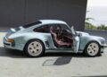 A light blue classic sports car, part of the California-Based Singer’s latest reimagined Porsche 911 Carrera Coupes unveiled at Goodwood, sits with its driver's door open to reveal a pink interior in a modern, oceanside setting.