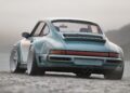 A classic blue sports car with wide tires, part of the California-Based Singer’s latest reimagined Porsche 911 Carrera Coupes, is parked on a rocky surface near water, viewed from the rear amid a misty, mountainous background.