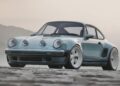 A vintage-style blue coupe with wide fenders and three round lights on the hood, reminiscent of the California-Based Singer Unveils Latest Reimagined Porsche 911 Carrera Coupes at Goodwood, is parked on a flat, sandy surface with hills in the background.