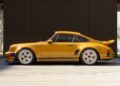 A yellow classic sports car, reimagined by a California-based singer, is parked indoors in side view, sunlight and trees visible outside through large openings—one of the latest Porsche 911 Carrera Coupes unveiled at Goodwood.