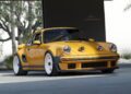 A yellow classic sports car with white wheels is parked under a modern structure, as a California-based singer unveils the latest reimagined Porsche 911 Carrera Coupes at Goodwood, with trees and greenery in the background.