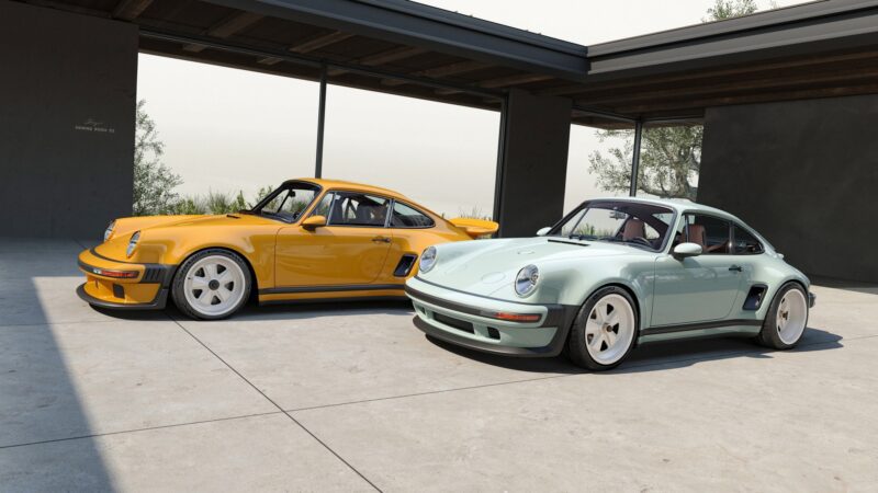 Two vintage sports cars, including a California-based singer's latest reimagined Porsche 911 Carrera Coupes, are parked side by side in a modern, open garage with large glass walls and a concrete floor.