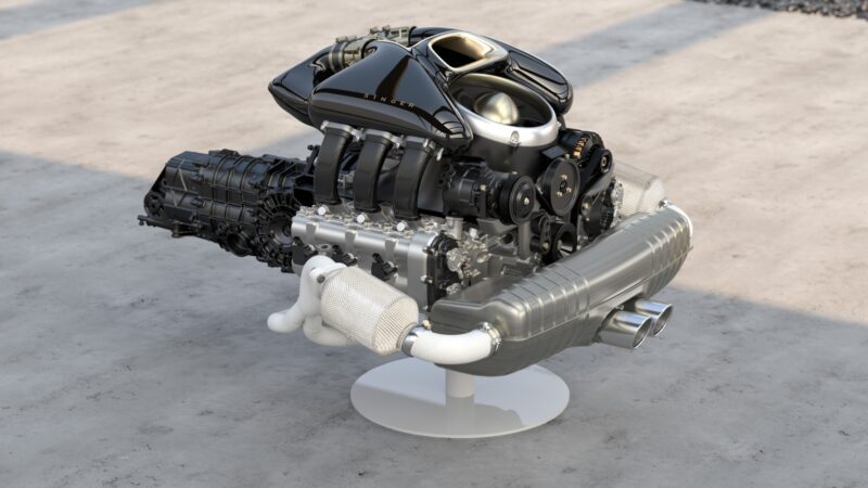 A high-performance automotive engine with visible intake system, exhaust manifold, transmission, and exhaust pipe displayed on a stand, similar to those powering California-based Singer’s latest reimagined Porsche 911 Carrera Coupes unveiled at Goodwood.