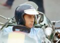 A man wearing a helmet sits in the cockpit of a vintage race car, looking to the side, as Lotus prepares to honor F1 legends Senna and Clark at Goodwood; part of another person is visible in the background.
