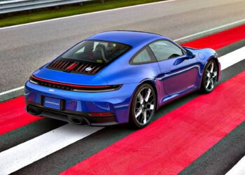 A blue Limited-Run 911 Carrera T Club Coupe sports car is parked on a red and white striped section of a racetrack, viewed from the rear left side.