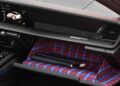 The open glove compartment of a Limited-Run 2026 911 Carrera T Club Coupe features a blue and red plaid interior lining, a black folder, and a charging cable; the dashboard displays a digital screen.