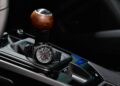 A black wristwatch rests on the leather boot of a car's manual gear shift knob—its wood and metal finish evokes the craftsmanship of the Limited-Run 2026 911 Carrera T Club Coupe, celebrating 70 years of Porsche Club of America.
