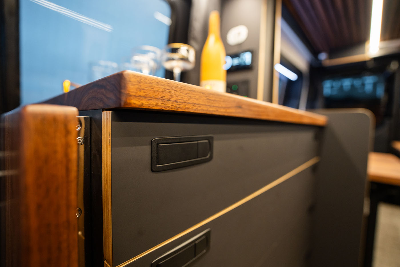 Close-up of a modern kitchen counter with a wooden edge and matte drawers; bottles and glasses are visible in the background.