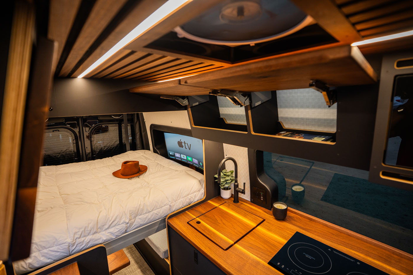 Modern camper van interior featuring a bed with a hat, mounted TV, wooden cabinets, kitchenette with stove, sink, and potted plant.