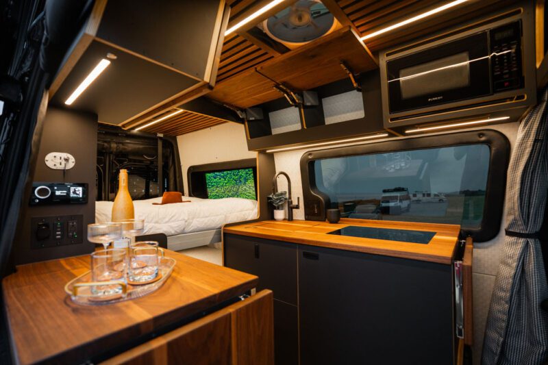 Modern camper van interior with wooden cabinetry, a kitchen area with sink, glassware on a counter, a microwave, and a bed in the background.