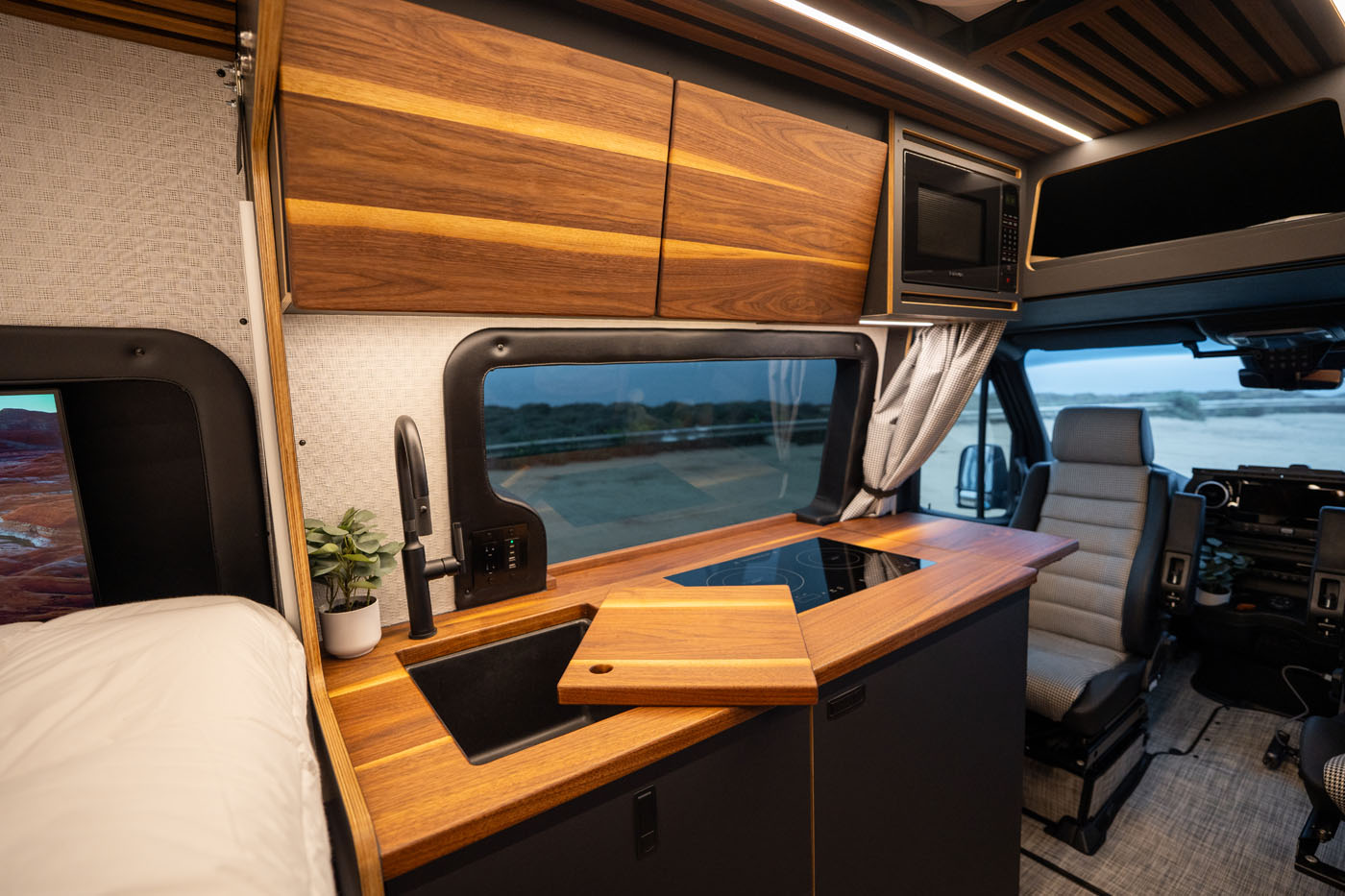 Modern camper van interior with wood cabinetry, a countertop with a built-in stove and sink, potted plant, and a view out the window toward an outdoor landscape.