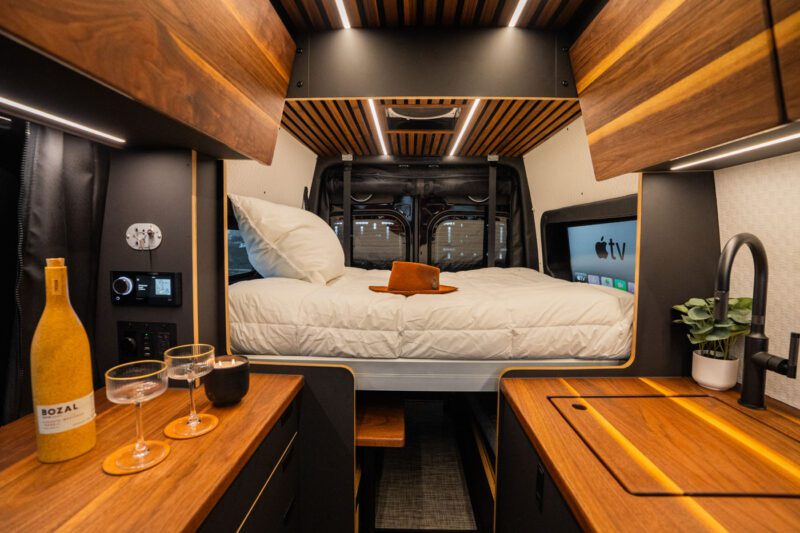 Interior of a modern camper van featuring a neatly made bed, wooden cabinets, a countertop with wine and glasses, plants, and ambient lighting.