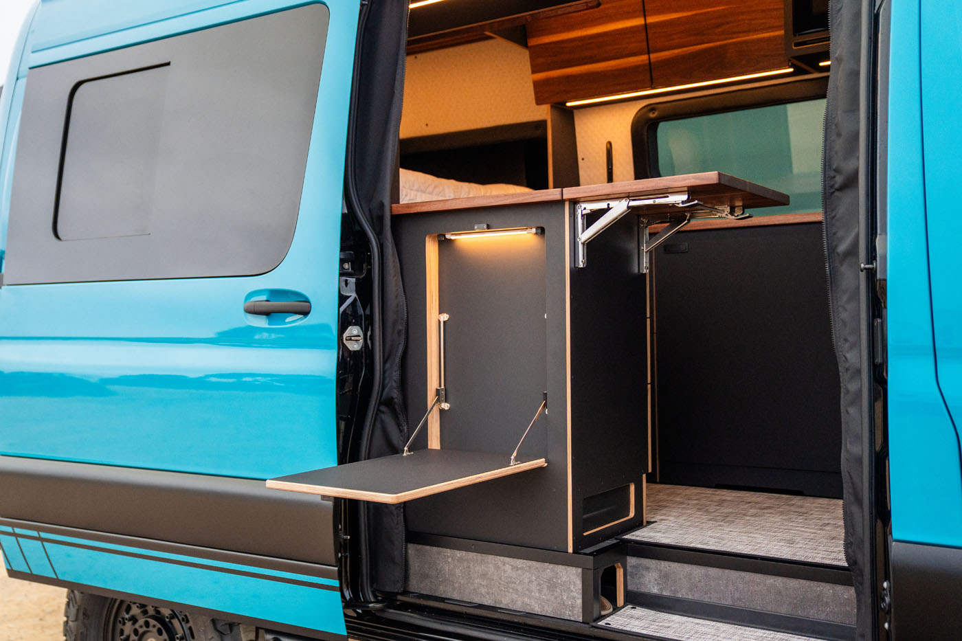 A blue camper van with its sliding door open reveals a compact interior featuring a fold-out table, wood cabinets, and modern lighting.