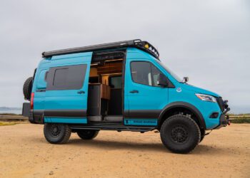 A blue camper van with off-road tires is parked on sandy ground with its side door open, revealing a partial view of the interior.