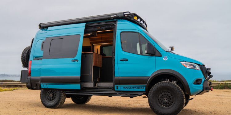 A blue camper van with off-road tires is parked on sandy ground with its side door open, revealing a partial view of the interior.