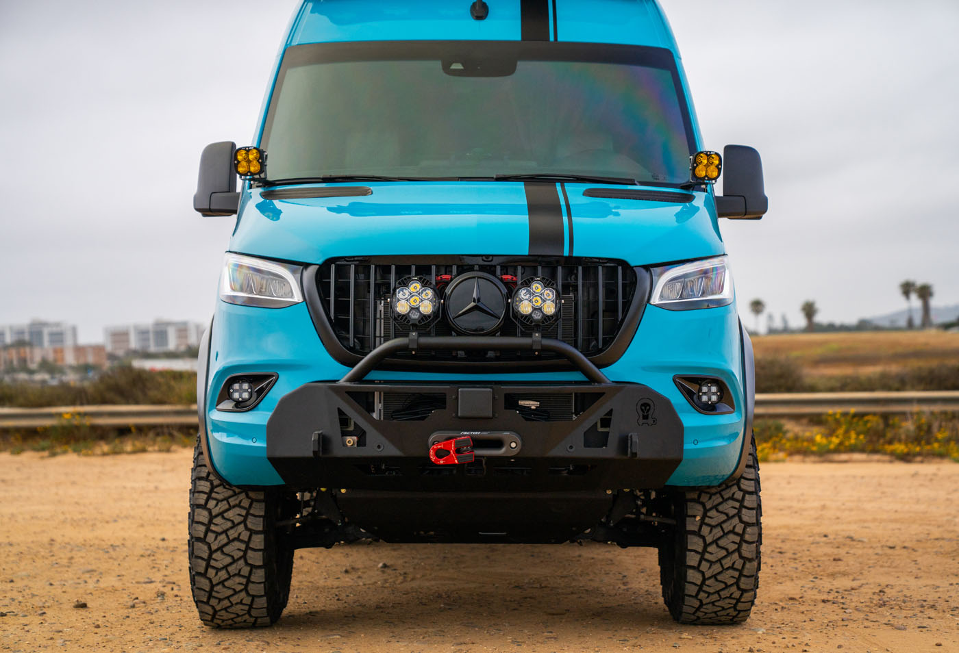 A blue Mercedes-Benz Sprinter van with off-road tires, extra lights, and a front bumper winch is parked outdoors on a dirt surface.
