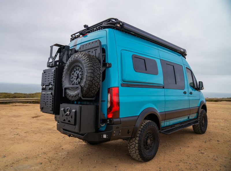 A blue off-road camper van with rugged tires, roof rack, and rear-mounted spare tire is parked on a dirt lot under an overcast sky.