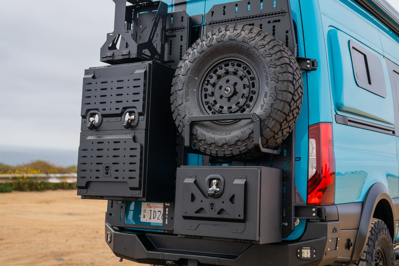 Back of a blue camper van equipped with a mounted spare tire and two black metal storage boxes on a dirt road.