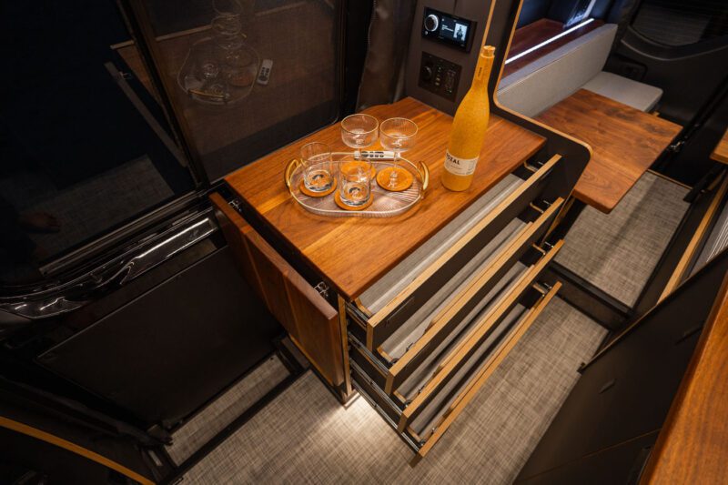 A wooden pull-out counter in a compact kitchen holds a bottle, four glasses, and a tray; several drawers are open below the counter.