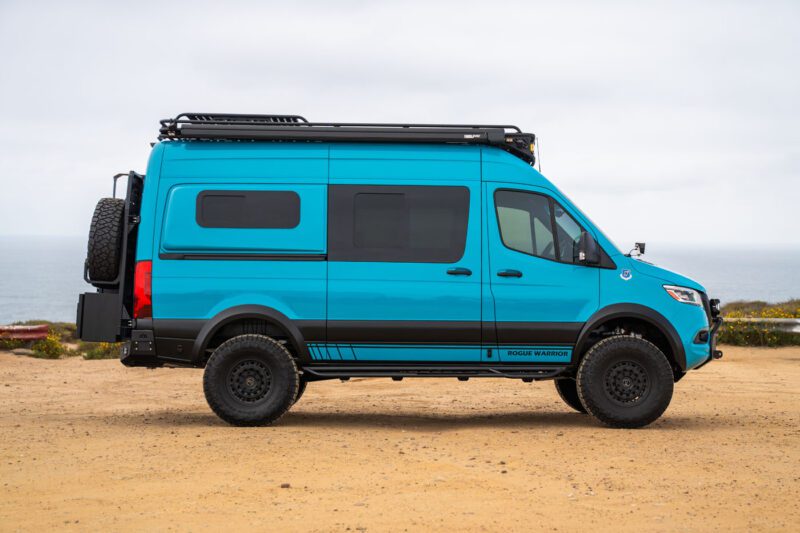 A blue camper van with off-road tires and roof rack is parked on a sandy surface near the coast under an overcast sky.