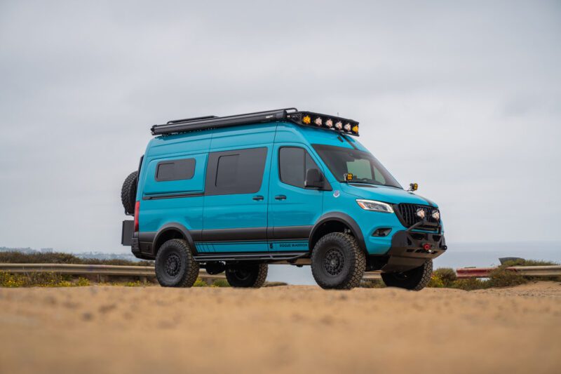 A blue camper van with off-road tires and roof rack is parked on a sandy area near the coast under a cloudy sky.