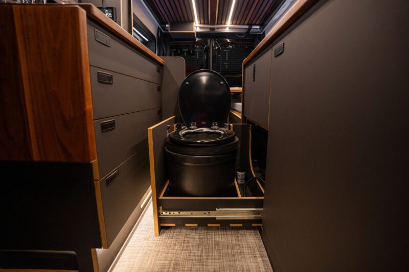 A compact composting toilet with the lid open is built into a pull-out drawer between cabinets inside a modern van conversion.