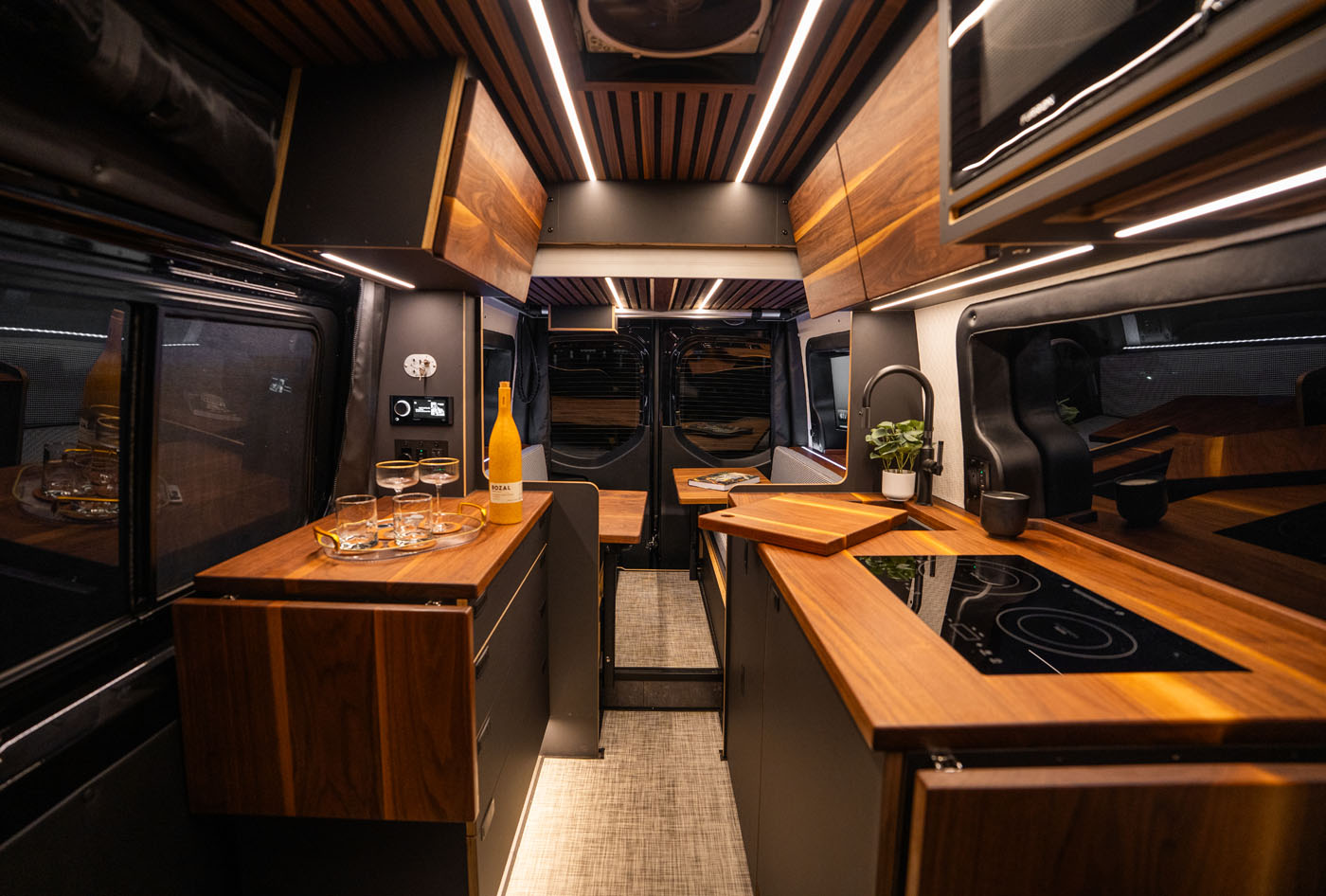 Modern camper van interior with wood cabinetry, built-in stove, sink, and a tray with glasses and a bottle on the counter, illuminated by overhead lights.