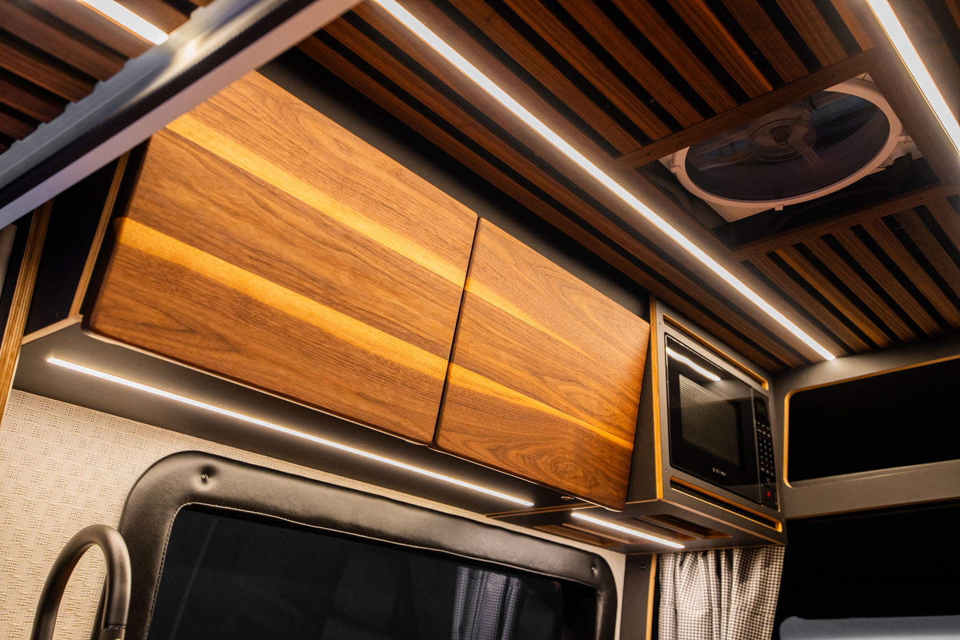 Wooden overhead cabinets, a microwave, and built-in LED strip lighting in the ceiling of a modern camper van interior.