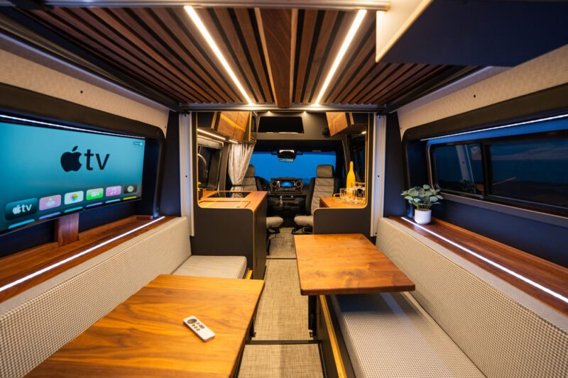 Interior of a modern camper van with wooden tables, cushioned benches, a TV displaying Apple TV, a remote, a plant, and a view toward the driver's seat.
