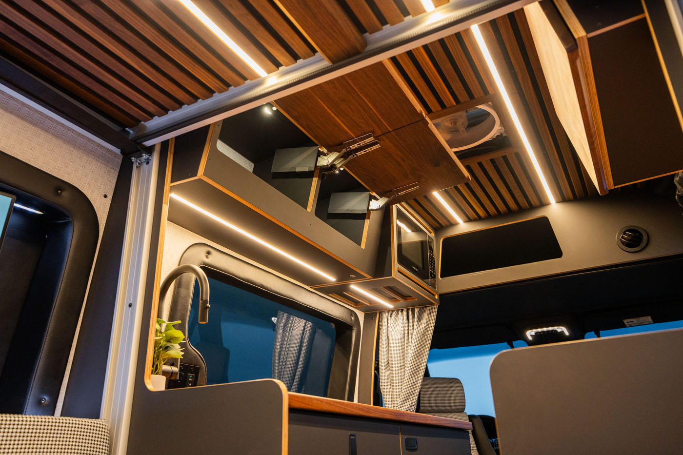 Modern camper van interior featuring wood paneling, built-in cabinets, LED lighting, a small sink, and a window with curtains.