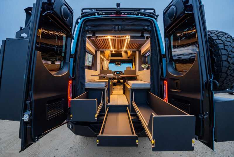 View of a camper van’s open rear doors showing interior seating, overhead lights, and extended storage drawers on a sandy surface.