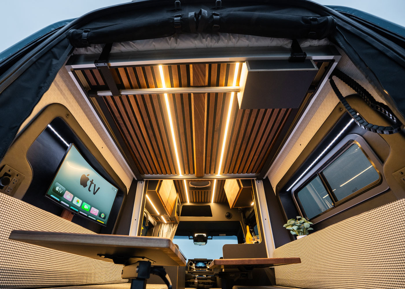 Interior of a modern camper van with wood panel ceiling, LED lights, a TV displaying Apple TV, a small table, and seating area.