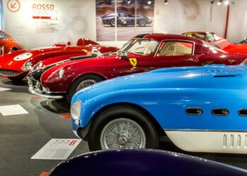 A row of classic sports cars, including a blue and several red models, displayed in a well-lit automotive museum exhibit.