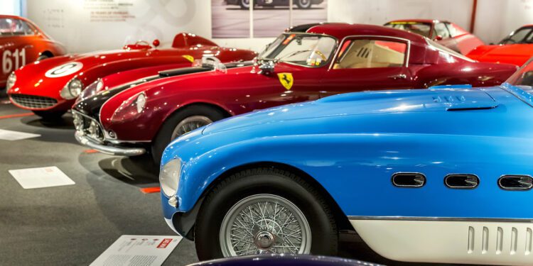 A row of classic sports cars, including a blue and several red models, displayed in a well-lit automotive museum exhibit.