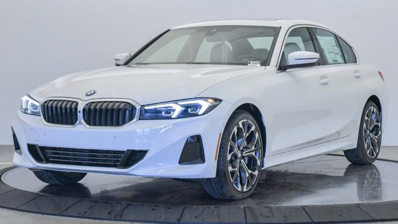 A white BMW sedan, available with the best lease deals, is parked indoors on a rotating platform, facing left with headlights on and window stickers visible inside.