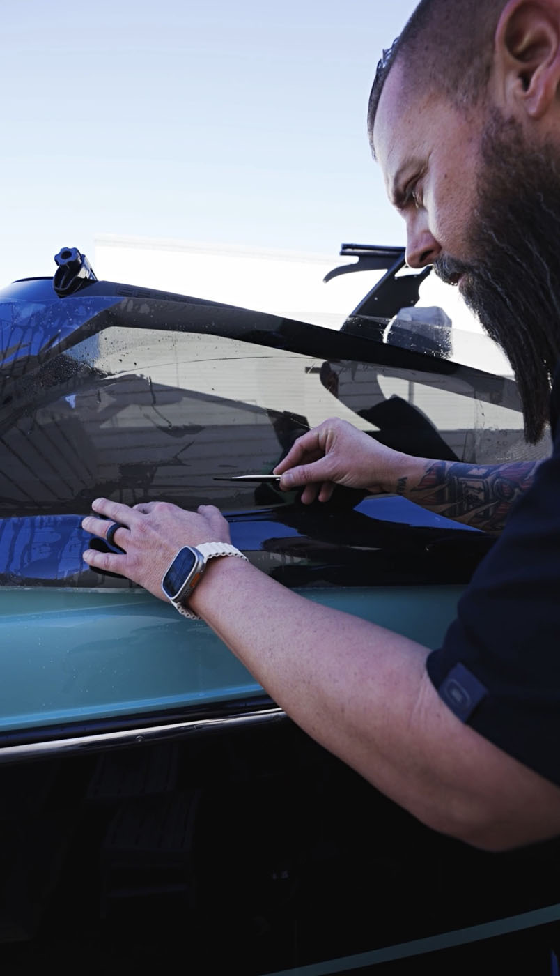 A person applies black vinyl wrap to the side of a vehicle, smoothing it out with a tool while wearing a smartwatch and a ring, demonstrating how protective films can enhance both style and durability.