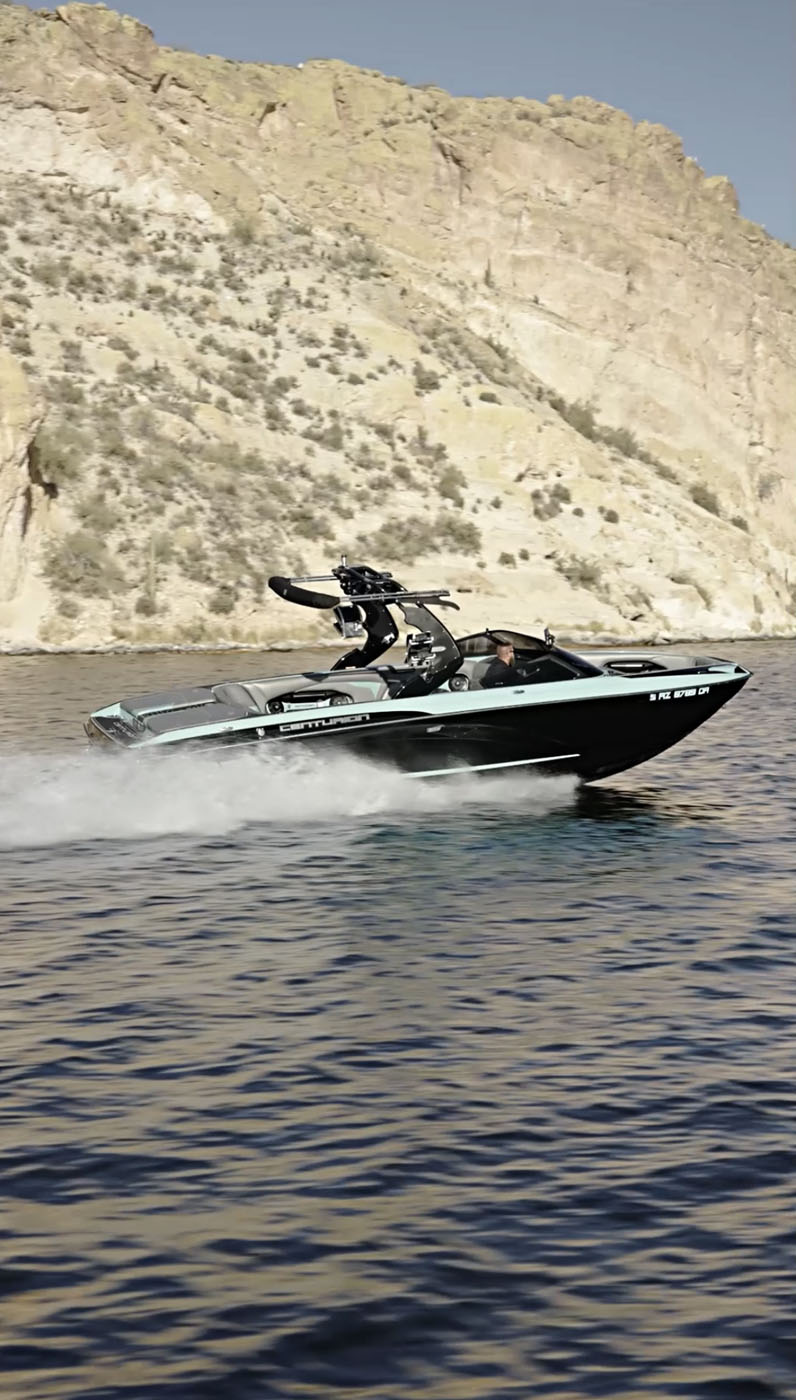 A black speedboat with protection coatings moves quickly across a lake near rocky cliffs, creating a wake behind it under clear skies.