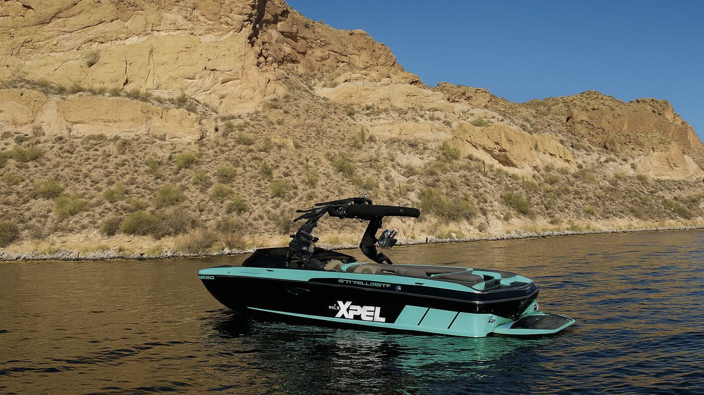 A modern speedboat with "XPEL" branding, featuring advanced protection coatings, floats on calm water near a rocky desert shoreline under a clear blue sky.