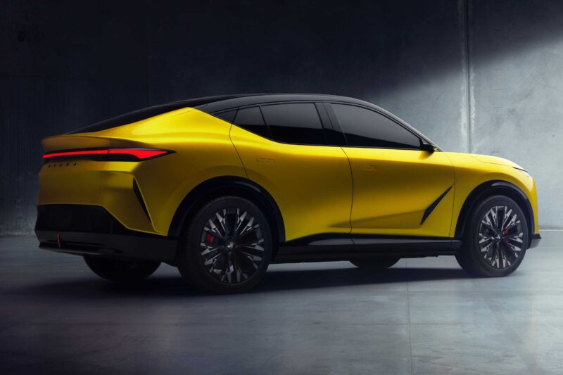 A yellow, modern all-electric SUV with tinted windows and black roof is parked indoors on a concrete floor under dramatic lighting, reminiscent of a reveal in Monterey.