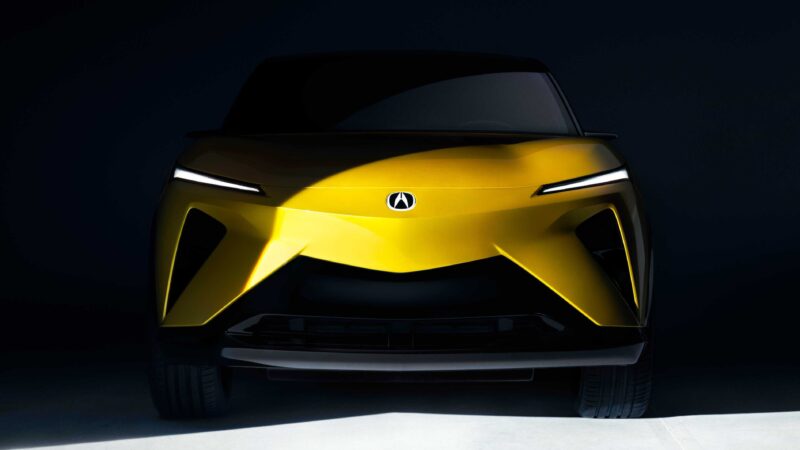 Front view of a yellow Acura RSX concept car with narrow headlights, a bold angular design, and the Acura logo centered on the hood, set against a dark background at Monterey.