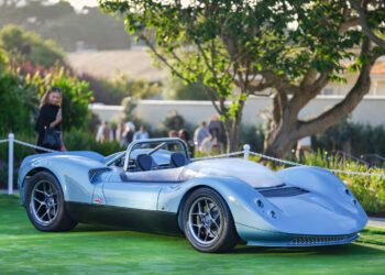 A sleek, light blue vintage-styled sports car is displayed on a green lawn with trees and people blurred in the background.