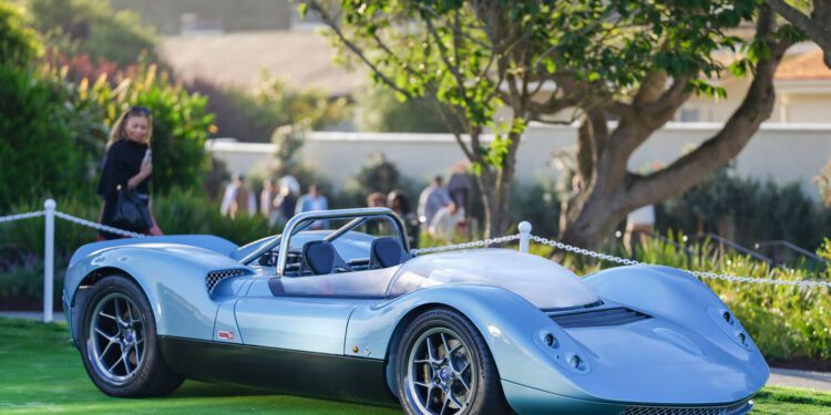 A sleek, light blue vintage-styled sports car is displayed on a green lawn with trees and people blurred in the background.