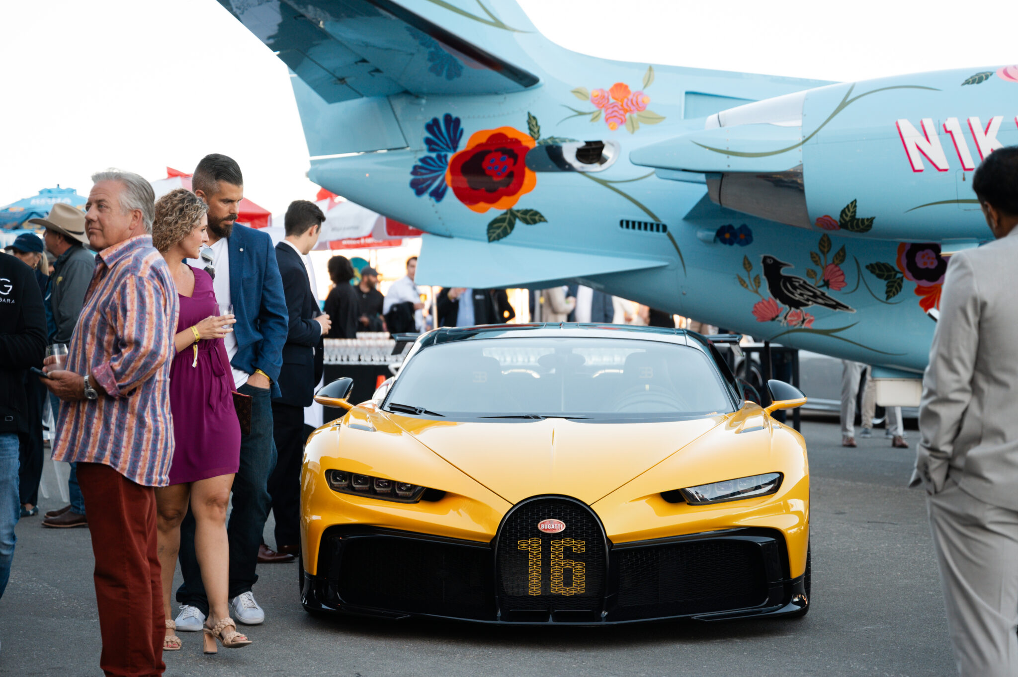 A yellow Bugatti sports car is parked in front of a blue aircraft with floral designs at Monterey Car Week 2025, as people stand and converse nearby—a highlight showcased by duPont REGISTRY.
