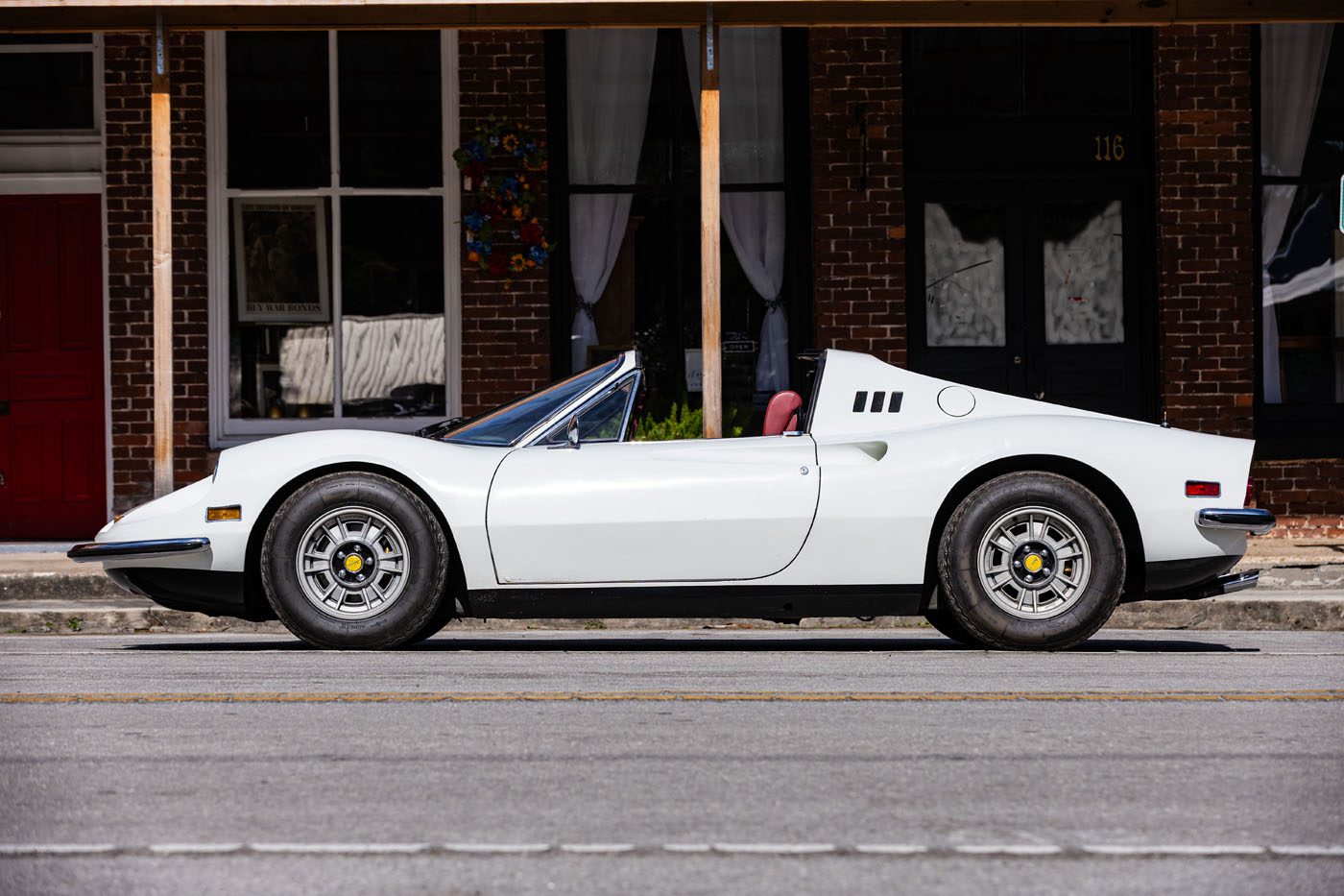 A white classic sports car with a convertible top, reminiscent of Ferrari Legends, is parked on the street in front of a brick building—an icon you might spot at the Monterey auction.