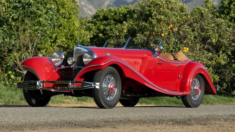 A red vintage convertible car with chrome details and tan interior is parked on a road, surrounded by greenery and mountains—an iconic sight reminiscent of the 2025 RM Sotheby's Monterey Auction: Top 10 Results and Highlights.