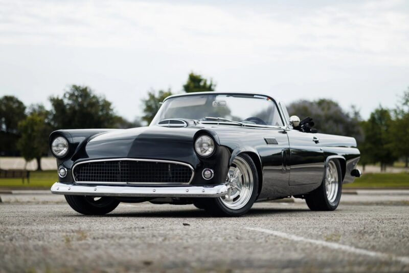 A classic black convertible muscle car is parked on an empty lot, with trees and a cloudy sky in the background—a must-see for fans of classic muscle cars.
