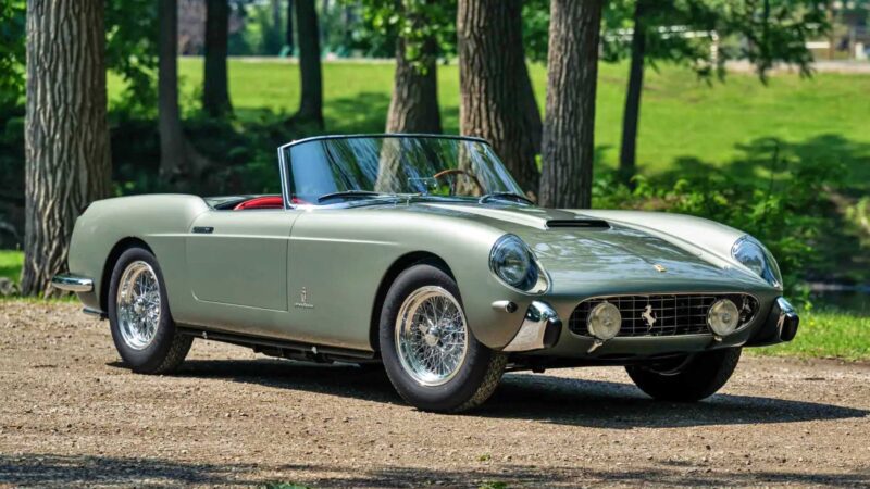 A classic silver convertible sports car with wire-spoke wheels is parked on a dirt path in a wooded, green park setting—a scene reminiscent of the elegance seen at the 2025 RM Sotheby's Monterey Auction: Top 10 Results And Highlights.